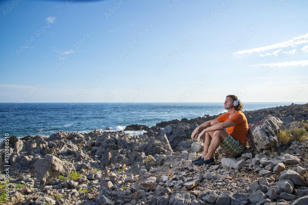 Adult man relaxing on a rocky cliff, sitting and looking at sea and mountains, Summer time Concept of freedom