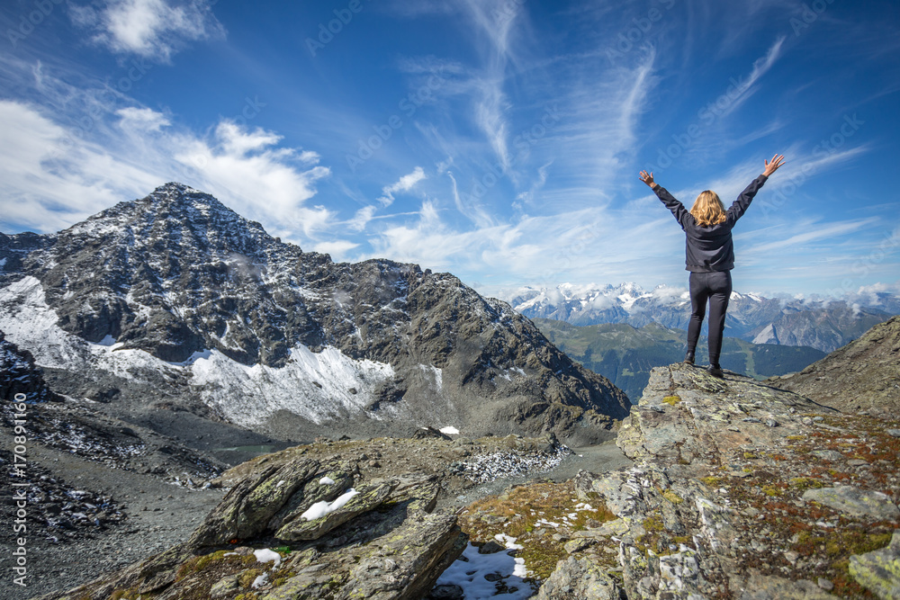 une jeune fille blonde debout , les bras en l'air  face à un paysage de montagne avec des cimes enneigées