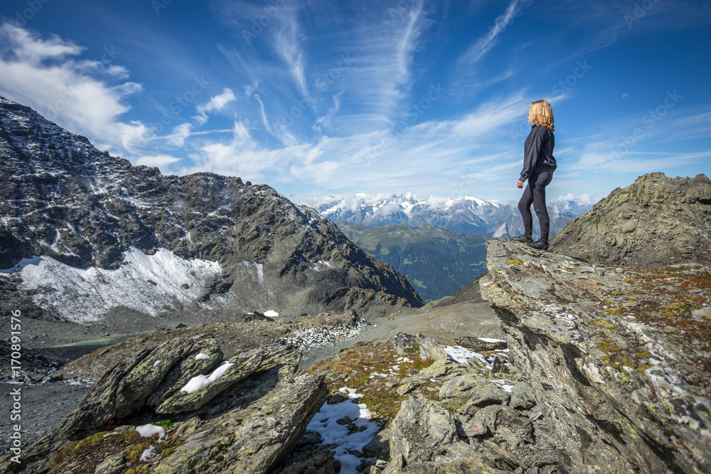 une jeune fille blonde debout et de dos admire un paysage de montagne avec des cimes enneigées