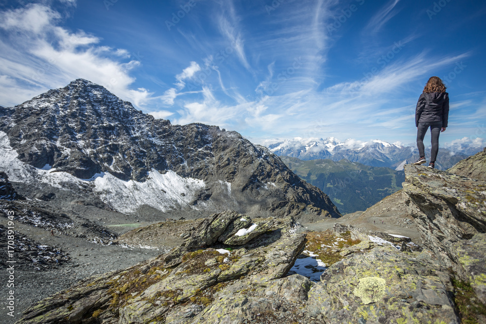 une jeune femme brune debout et de dos admire un paysage de montagne avec des cimes enneigées