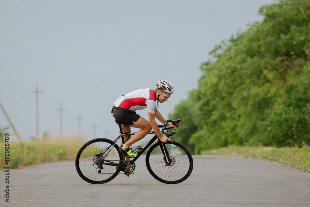 Obraz premium Bicycle racer in helmet and sportswear training alone on empty country road, fields and trees background 