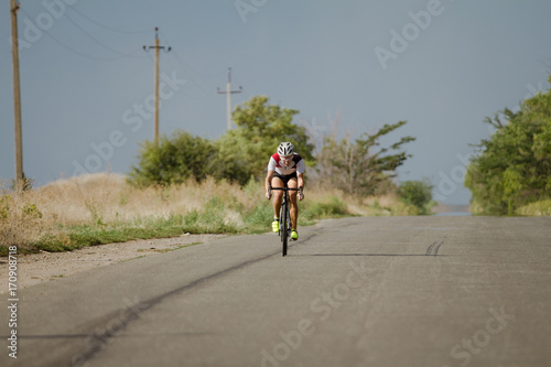 Bicycle racer in helmet and sportswear training alone on empty country road  fields and trees background   