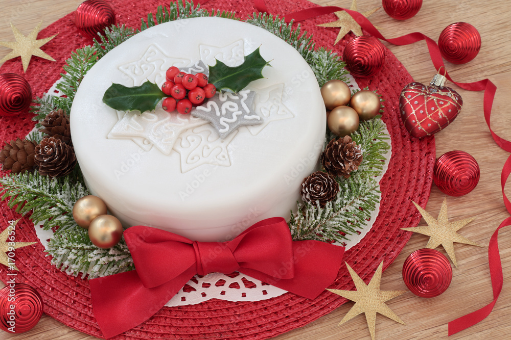 Iced christmas cake with red ribbon and bow, holly, fir and bauble decorations on oak table background.