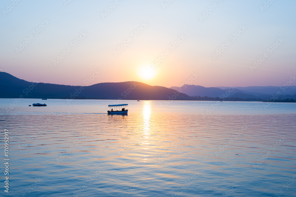 Boats floating on Lake Pichola with colorful sunset reflated on water beyong the hills. Udaipur, Rajasthan, India.