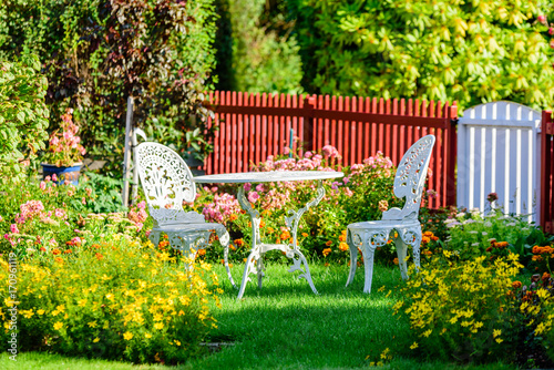 White metal garden furniture among flowers on a lawn. Picket fence in background.