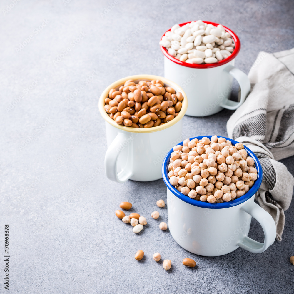 Raw uncooked organic chickpeas and kidney beans in white enamel mugs on rustic wooden board on light gray stone background. Healthy vegan vegetarian food concept with copy space. Raw uncooked organic chickpeas and kidney beans in white enamel mugs on rustic wooden board on light gray stone background. Healthy vegan vegetarian food concept with copy space.