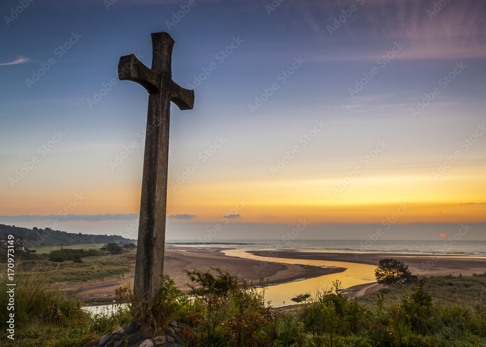 The Wooden Cross on Church Hill, Alnmouth, Northumberland, England. UK. The hill is the site of the long ruined church of St Waleric. At sunrise/ dawn with a colourful dawn sky.