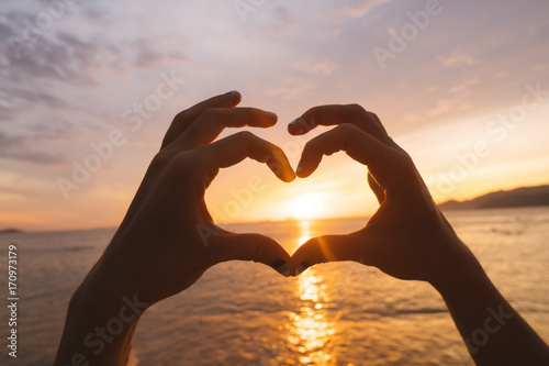 Hands and fingers in heart shape framing setting sun at sunrise over ocean