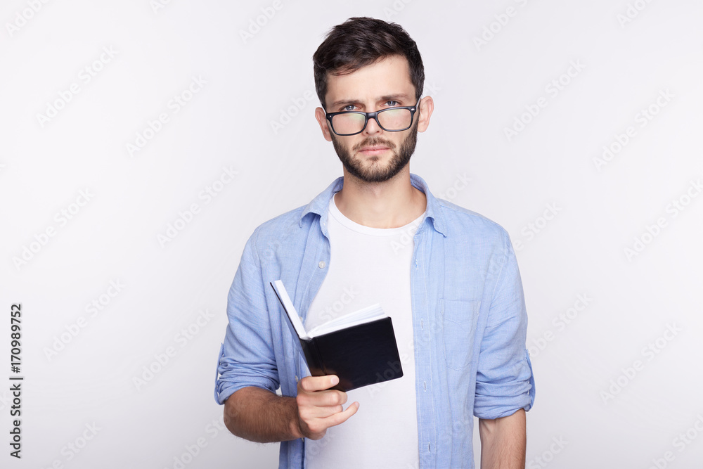 Casually dressed young handsome male bearded student with blue eyes wearing glasses has focused concentrated look as he reads information in textbook and making notes in copybook, preparing for exam.
