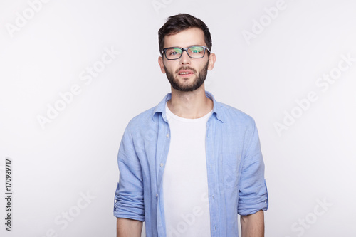 Caucasian young hipster wearing glasses in black frame and blue shirt looking at camera with indifferent stony facial expression, feeling confident and handsome against posing white studio wall. © Yulia