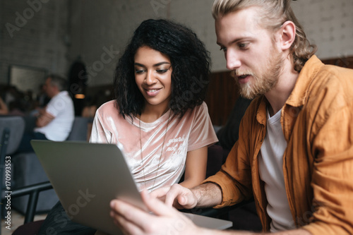 Portrait of young man and girl sitting in restaurant and working on laptop. Smiling African American girl with dark curly hair sitting at cafe with friend that holding laptop in hands