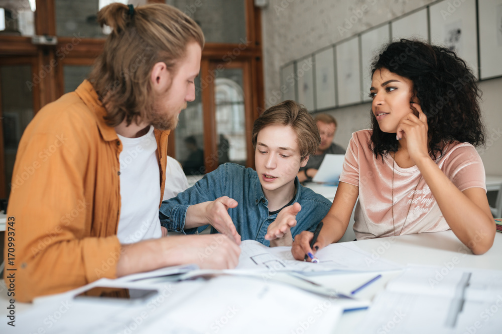 Portrait of young people emotionally discussing something in office. Two boys with blond hair and girl with dark curly hair sitting and working together in classroom. Students studying together Portrait of young people emotionally discussing something in office. Two boys with blond hair and girl with dark curly hair sitting and working together in classroom. Students studying together