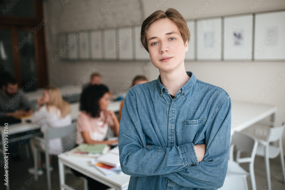 Fototapeta premium Portrait of young man with blond hair looking in camera in office. Cool boy in denim shirt standing with clasped hands in classroom with students on background