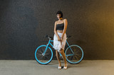 Pretty young trendy woman posing with bicycle next to modern black stone wall 