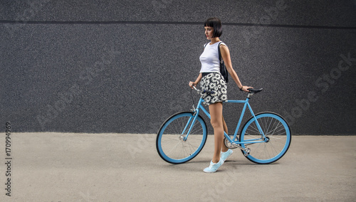Urban hipster girl with all blue bicycle next to modern wall, copy space 
