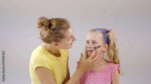 smiling woman is applying stripes of dark clay on a face of her little blond-haired daughter photo