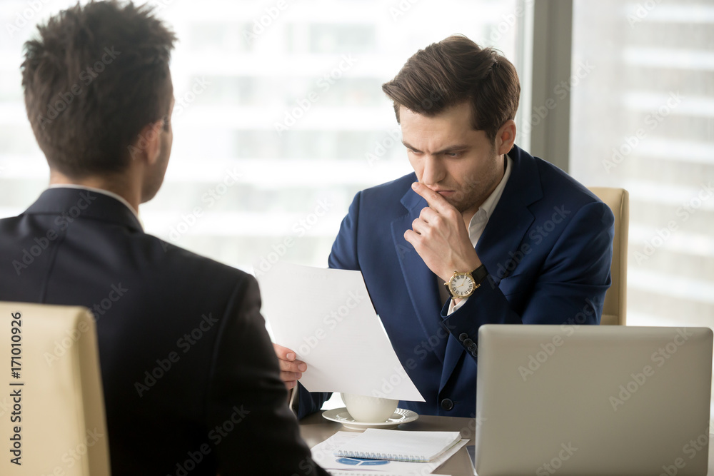 Doubting businessman reading contract document with suspicion when sitting at desk in front of business partner. Entrepreneur unsure in terms of agreement, feeling skeptical because of financial plan Doubting businessman reading contract document with suspicion when sitting at desk in front of business partner. Entrepreneur unsure in terms of agreement, feeling skeptical because of financial plan
