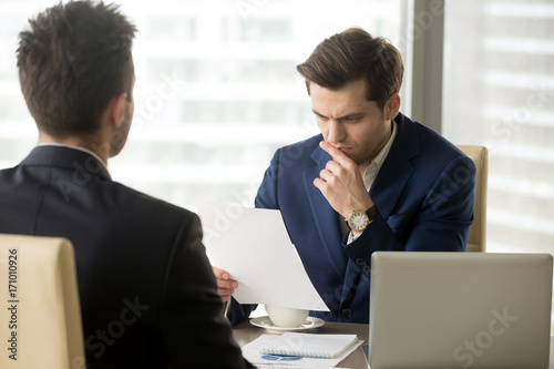 Doubting businessman reading contract document with suspicion when sitting at desk in front of business partner. Entrepreneur unsure in terms of agreement, feeling skeptical because of financial plan
