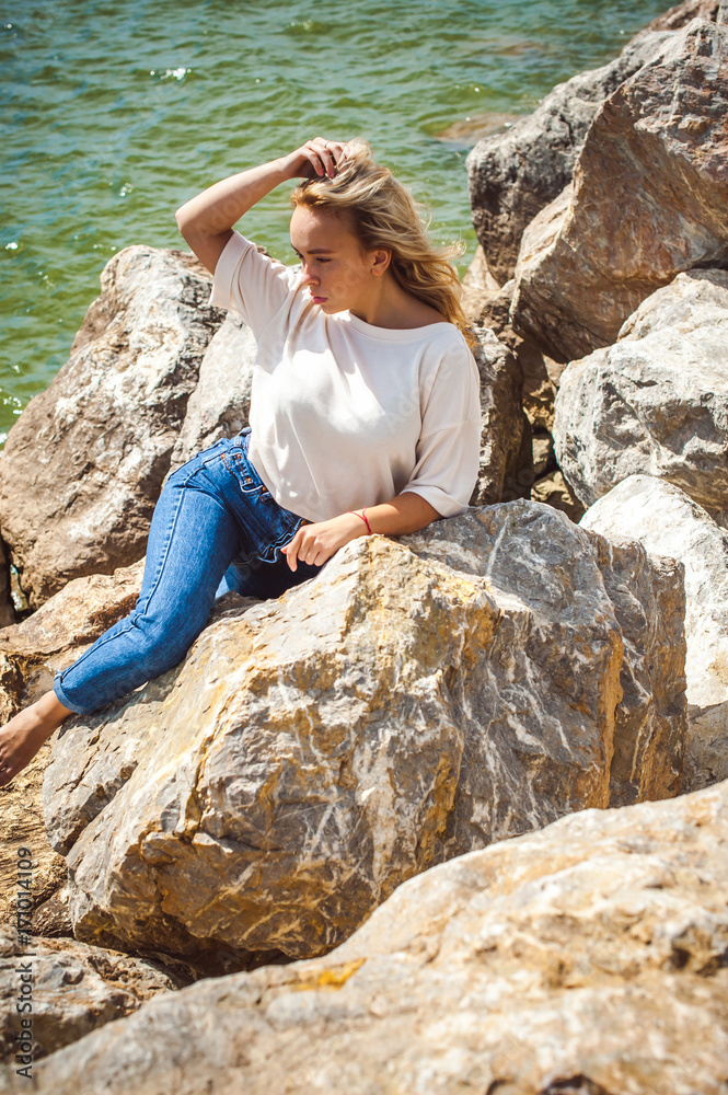 Portrait young woman on stones near sea. Beautiful girl is resting on coast, enjoying outdoor recreation.