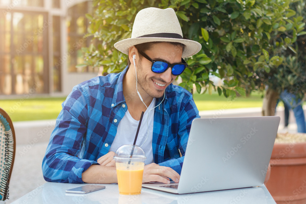 Closeup photo of young handsome Caucasian male looking at screen of his laptop and listening to voice through earphones with excitement while eating out in outdoor street cafe, smiling happily