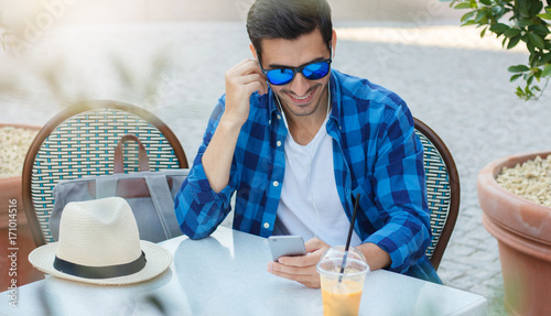Horizontal summer photo of handsome European man in casual clothes spending afternoon in outdoor cafe while communicating with dearest and nearest on phone via headphones sharing positive content