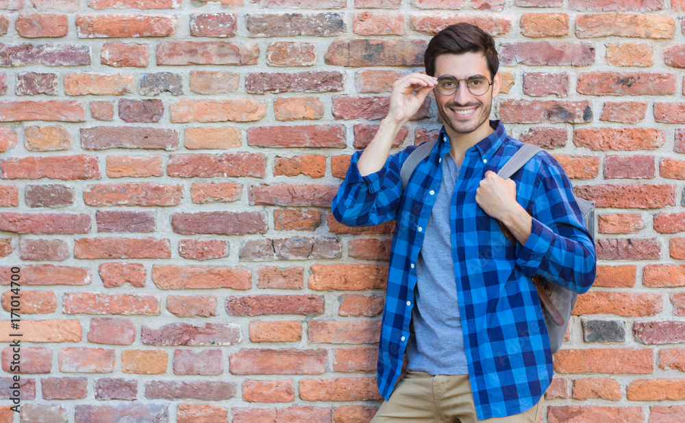 Horizontal image of sporty European man pictured with red brick wall behind ready for discovering new places with backpack on his back and fashionable black-rimmed spectacles lit with daylight Horizontal image of sporty European man pictured with red brick wall behind ready for discovering new places with backpack on his back and fashionable black-rimmed spectacles lit with daylight