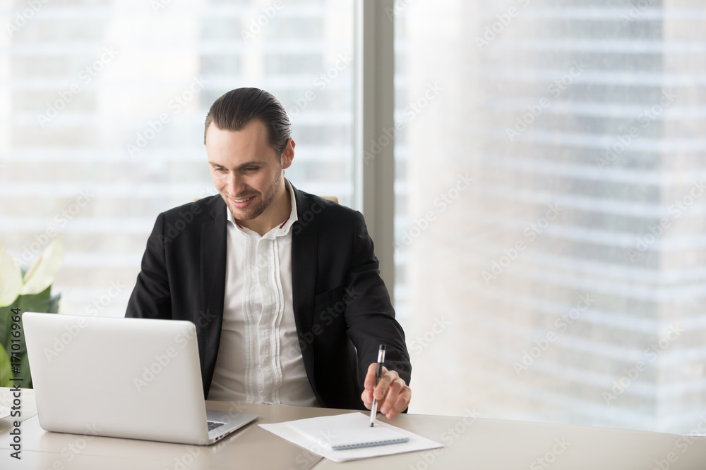 Young cheerful business man sitting in modern office, looking at laptop with happy smile, pleased with successful deal, financial report, project results and stock investment profit. Success concept. Young cheerful business man sitting in modern office, looking at laptop with happy smile, pleased with successful deal, financial report, project results and stock investment profit. Success concept.