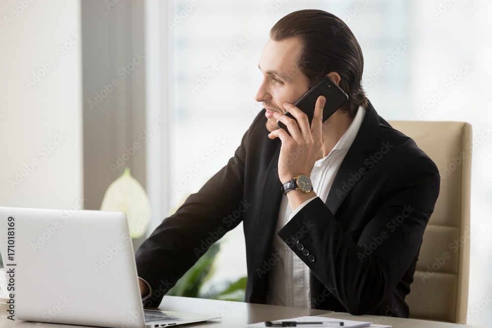 Smiling young businessman in modern office at work desk in front of laptop talking on cellphone. Successful deal discussion, planning future meetings, business negotiations, client consulting concept. Smiling young businessman in modern office at work desk in front of laptop talking on cellphone. Successful deal discussion, planning future meetings, business negotiations, client consulting concept.