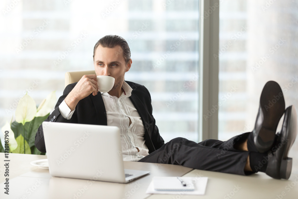 Businessman relaxing with legs up on desk, drinking coffee while dreaming about great future opportunities at workplace in modern office. Successful project manager resting after positive day at work. Businessman relaxing with legs up on desk, drinking coffee while dreaming about great future opportunities at workplace in modern office. Successful project manager resting after positive day at work.