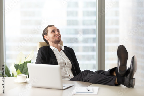 Happy smiling businessman relaxing, with legs up on desk at workplace in modern office. Dreaming about opportunities, taking break from workflow, chilling after good meeting, finished project concept.