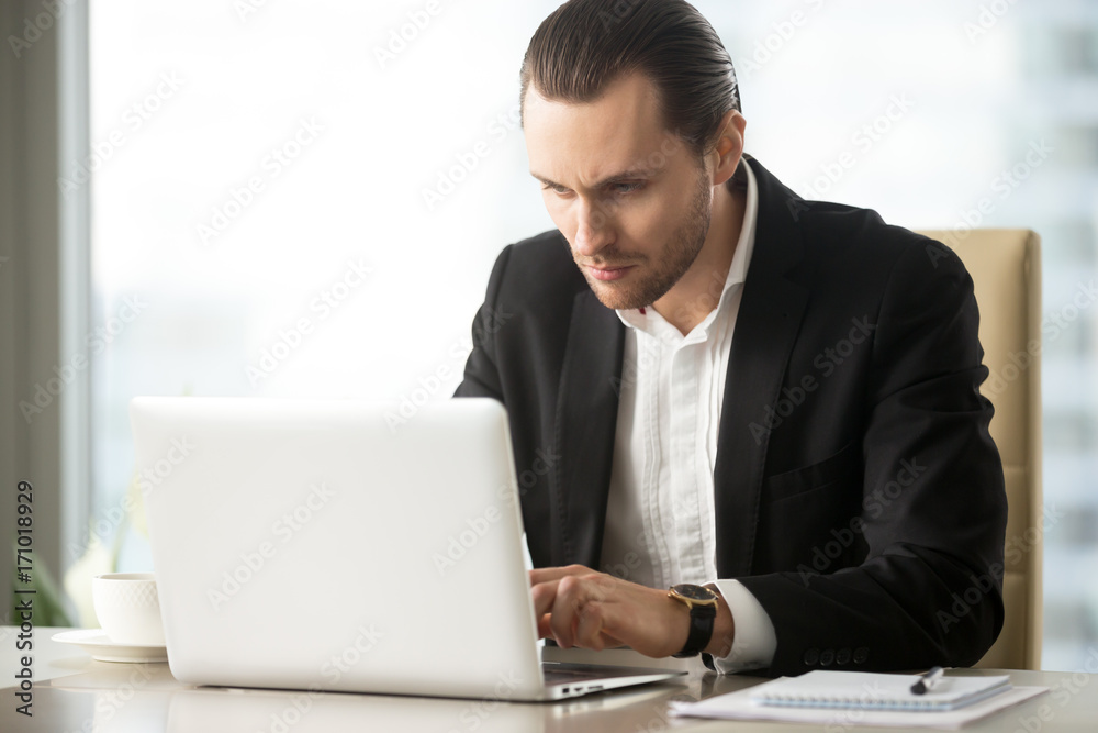 Serious young handsome busy businessman working on laptop at desk at workplace in office. Researching stock investment market, communicating with business partners, taking care of financial inquiries. Serious young handsome busy businessman working on laptop at desk at workplace in office. Researching stock investment market, communicating with business partners, taking care of financial inquiries.