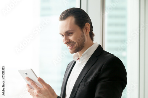 Smiling handsome businessman in office, looking at cellphone or electronic tablet with cheerful face. CEO happy about great stock investment news, remote communication with business partner concept.