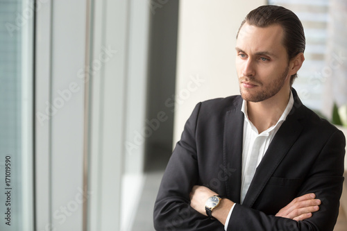 Serious young businessman standing at workplace with arms crossed on chest, in deep though about corporate future. Contemplating new investment, brainstorming about project, thinking about career.