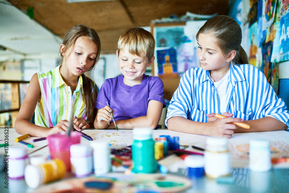 Naklejka premium Group of creative children having fun while sitting at desk and drawing with gouache, interior of art studio on background