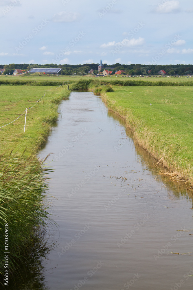 Dutch landscape with a broad ditch between meadows, in the distance a church tower and red tiled roofs of farmhouses