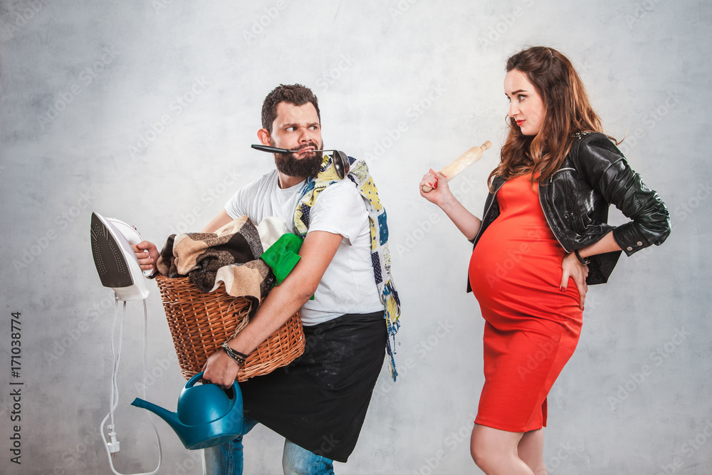 Foto Stock A beautiful pregnant woman with a big belly in a red dress with  a rolling pin in her hands grimacing at her husband who holds a basket with  clothes, iron