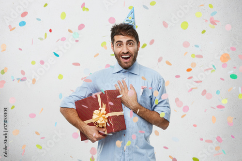 Photography Horizontal shot of excited happy adult male holding gift box and keeping hand on his chest, touched with sincere cordial toast made by friend on his birthday party