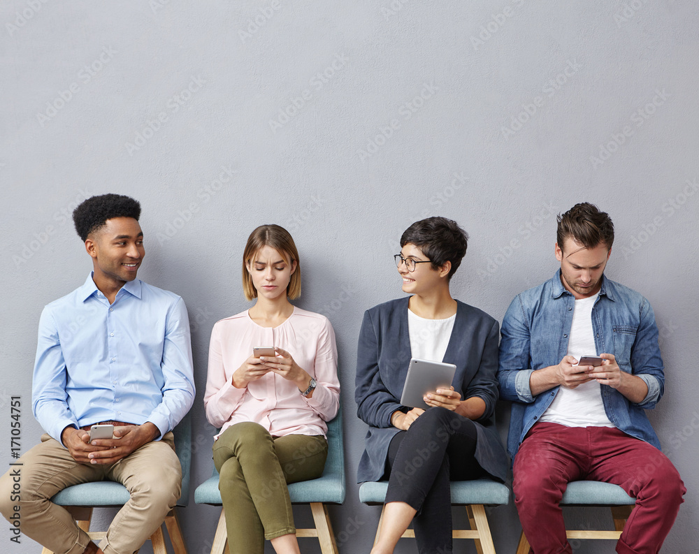 Multiethnic group of four beautiful young people in casual clothes using mobile phones while waiting for their turn in waiting room in bank, African man and woman in glasses talking to each other Multiethnic group of four beautiful young people in casual clothes using mobile phones while waiting for their turn in waiting room in bank, African man and woman in glasses talking to each other