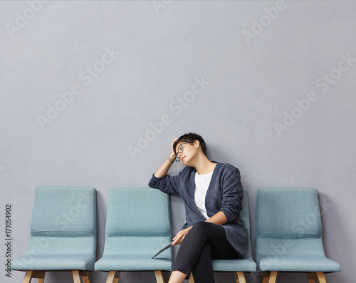 Portrait of attractive young mixed race female applicant for accountant position in large company feeling bored and tired while waiting for interview in hall, sitting on chair and using digital tablet