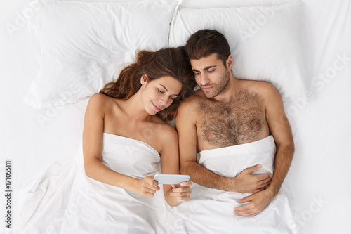 High angle view of beautiful Caucasian family male and female don't sleep at night lying in bed side by side surfing internet together, shopping online or browsing pictures using mobile phone photo