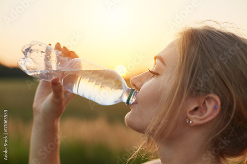 Fototapet Female athlete being thirsty after running, holding plastic bottle, drinking cold water, resting after jogging workout