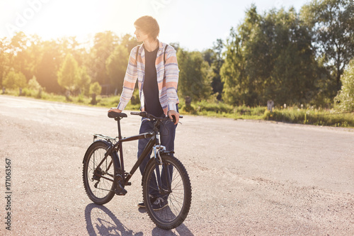 Hipster guy wearing shirt and jeans, standing with his bicycle, looking aside while waiting for his friend to come. Teenager going to school by bike. Handsome stylish boy cyclist. Healthy lifestyle
