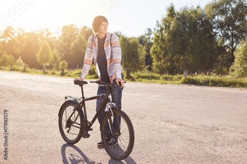 Pensive teenage boy standing on road, keeping hand on handle bar of his bike, waiting for others cyclists to have journey together, looking aside while standing on road. Active lifestyle concept