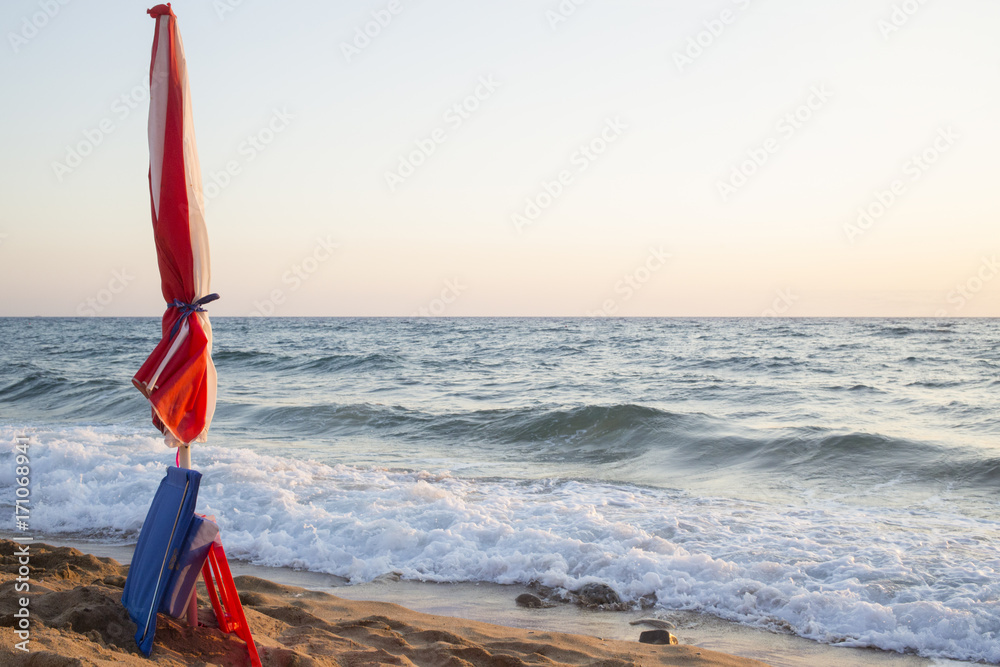 Dettaglio di un ombrellone chiuso su una spiaggia libera e vuota. Le onde  del mare agitato si alzano nell'ora del tramonto. L'ombrellone è rosso e  bianco a strisce. foto de Stock