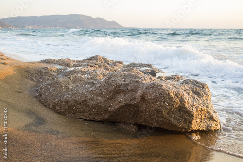 Dettaglio di uno scoglio di pietra che sta sulla spiaggia e viene bagnato dalle onde. L'acqua del mare ogni tanto arriva a riva e bagna la battigia della costa.