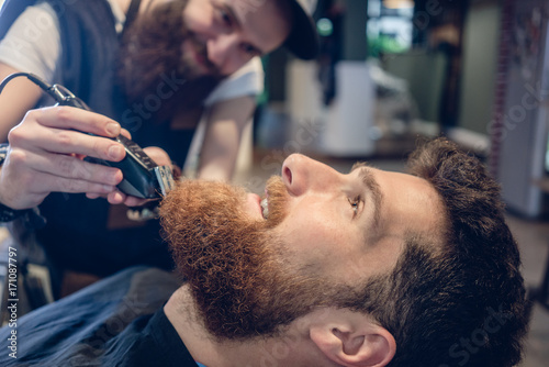 Side view close-up of the head of a redhead young man and the hand of a skilled barber, trimming his beard with an electric trimmer in a trendy hair salon for men