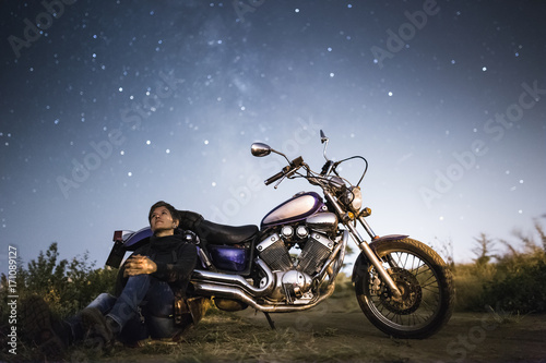 Fototapeta Naklejka Na Ścianę i Meble -  Young beautiful biker woman with motorcycle under stars of  home galaxy Miky Way. Female biker on field at night