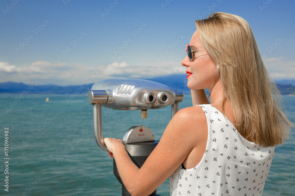 young sexy woman enjoy the beautiful view with a coin operated binoculars. The sun is shining, the water and the sky is blue. she wear sunglasses an a white dress