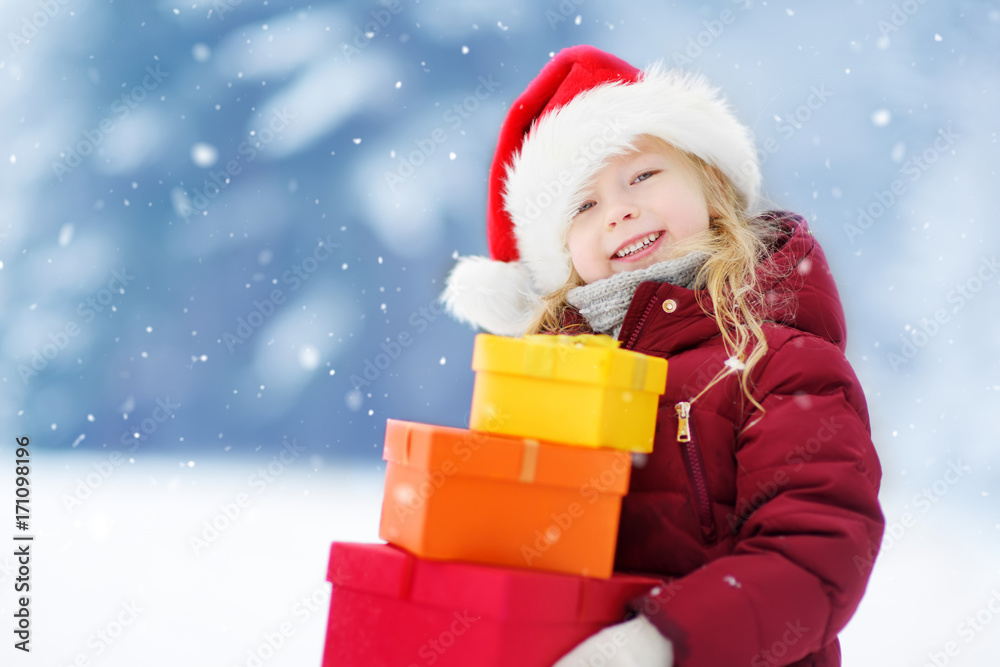 Adorable little girl wearing Santa hat holding a pile of Christmas gifts on beautiful winter day Adorable little girl wearing Santa hat holding a pile of Christmas gifts on beautiful winter day