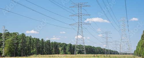 Parallel rows of transmission towers (power tower, electricity pylon, steel lattice tower) cloud blue sky in US. Texture high voltage pillar, overhead power line, industrial background. Panorama style
